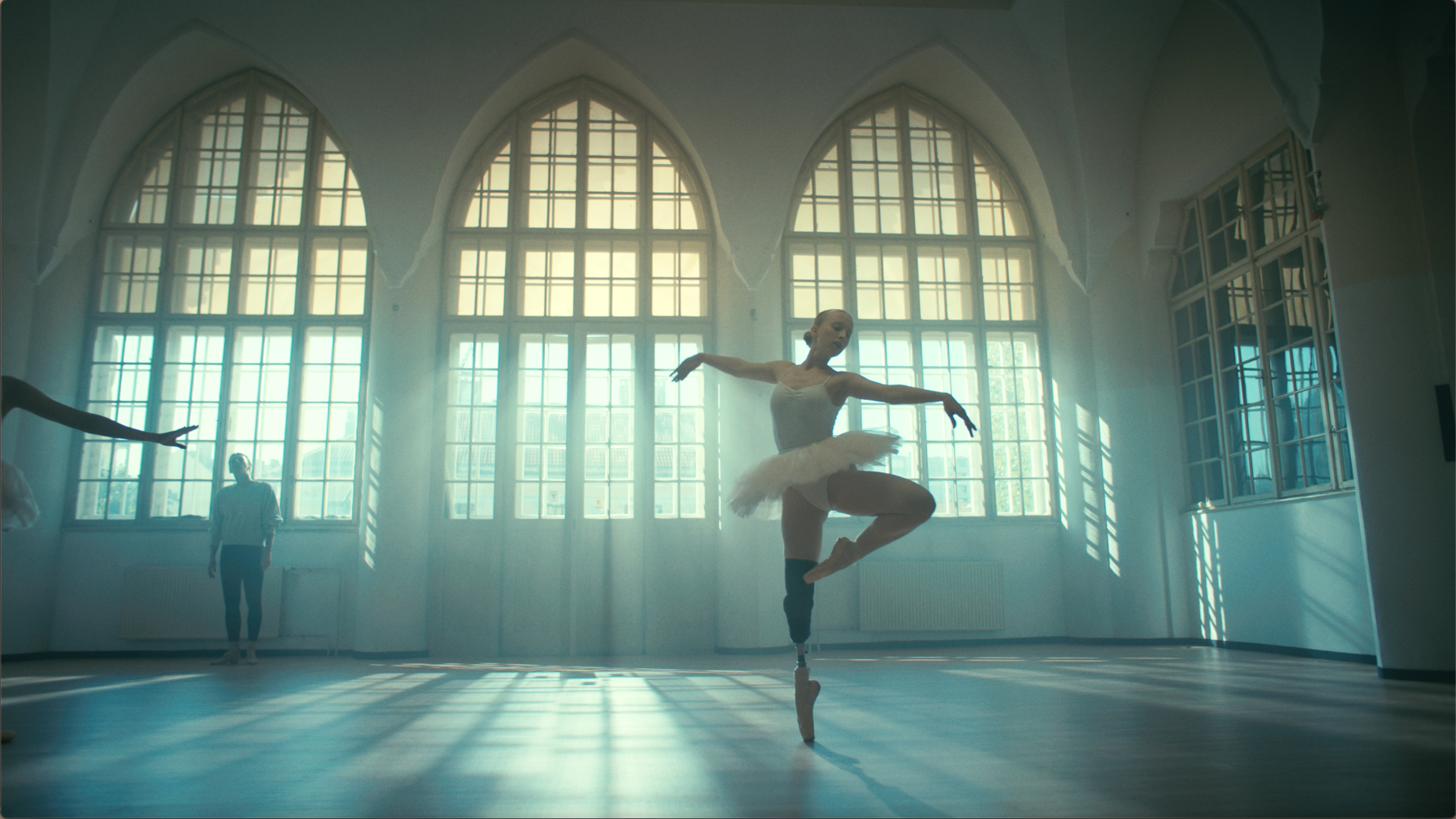A ballerina with a prosthetic leg is frozen in pose in a loft studio with rays of sunlight shining through large windows behind her.