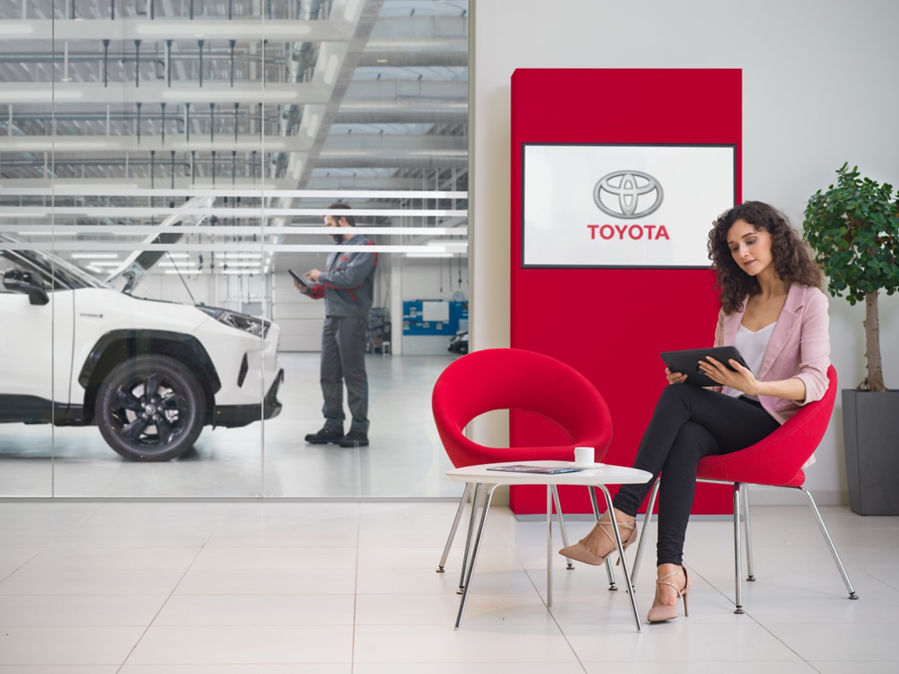 Een dame zit in een Toyota die wacht op onderhoud. Ze kijkt op een tablet, met op de achtergrond en TV-scherm waarop een logo van Toyota wordt weergegeven.