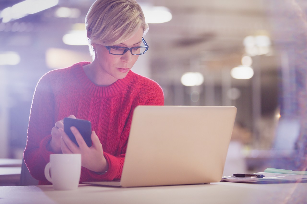 Een dame in een rode trui kijkt door haar bril op haar laptop, met in haar hand een smartphone. Op tafel staat een koffiekopje.